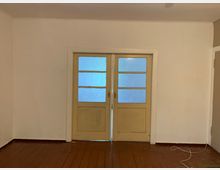 Ein einfacher Raum mit Holzboden und weißen Wänden, im Mittelpunkt eine zweiflügelige Glastür mit gelbem Holzrahmen und mattem Glas. Rechts unten liegt ein loses weißes Kabel auf dem Boden.