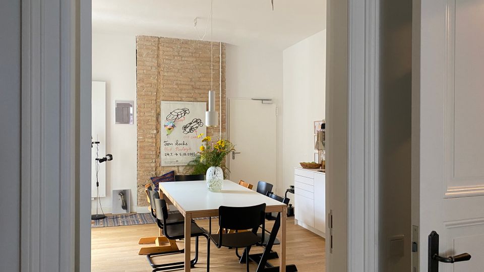 Blick durch einen Türrahmen in ein helles Esszimmer mit großem rechteckigem Tisch, schwarzen Stühlen und hängender Pendelleuchte. Im Hintergrund stehen eine freigelegte Ziegelwand mit Poster, ein Sideboard sowie eine Vase mit Blumen auf dem Tisch auf hellem Holzfußboden.