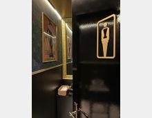 Schwarze Tür mit einem goldenen Piktogramm für eine Damentoilette, leicht geöffnet, Blick in einen dekorativen Innenraum mit goldgerahmtem Spiegel, Retro-Poster an der Wand und einem Korb mit Tüchern auf einer dunklen Ablage.