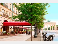 Straßenansicht des Hotels Adlon mit rotem Vordach und dekorativen Pflanzen am Eingang, flankiert von Bäumen. Im Hintergrund ist das Brandenburger Tor in Berlin sichtbar, links von parkenden Autos gesäumt.