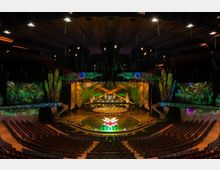 Ein großer, moderner Theatersaal mit einer aufwendig gestalteten Bühne, verziert mit Natur- und Pflanzenmotiven. Die Beleuchtung betont ein rundes Podium mit einer leuchtenden Blumenprojektion, während die Zuschauerreihen im Halbkreis um die Bühne angeordnet sind.