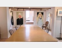 Ein heller Seminarraum mit einem langen Holztisch und schwarzen Metallstühlen. An den weißen Wänden hängen großformatige Gemälde, und durch ein Fenster fällt Tageslicht ein; im Hintergrund ist ein erhöhter Bereich mit einem weißen Sofa sichtbar.