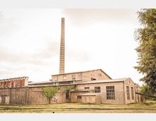 Backsteinfabrikgebäude mit hohem Schornstein, großen Fenstern und einem mit Efeu bewachsenen Teil der Fassade. Im Vordergrund befinden sich grasbewachsene Gleise, rechts Bäume.