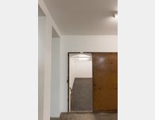 Ein schlichter Raum mit weißen Wänden und einem Boden aus dunklen Fliesen, gesehen durch eine geöffnete, rostfarbene Metalltür. Die Deckenbeleuchtung besteht aus mehreren rechteckigen Lampen.