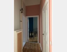 Ein schmaler Innenflur mit Holzboden, rosa und weiße Wände, sowie weißen Türen und Heizungsrohren an der linken Wand. Am Ende des Flurs ist ein Raum mit blauer Wand, Holzhocker und gestapelten Behältern sichtbar.