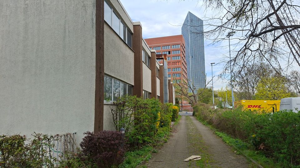 Schmaler gepflasterter Weg zwischen niedrigen Wohn- oder Bürogebäuden mit flachen Dächern und Sträuchern am Rand. Im Hintergrund ragen ein rotes Hochhaus und ein modernes Glashochhaus über Bäume und Straßenlampen hinweg.