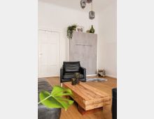 Modernes Wohnzimmer mit Holzboden, einem schwarzen Ledersessel und einem rechteckigen Couchtisch aus hellem Holz. Im Hintergrund stehen ein grauer Einbauschrank, dekoriert mit Pflanzen, Bildern und grünen Glasvasen, sowie eine weiße Tür.