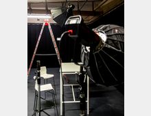 Ein Fotostudio mit schwarzen Vorhängen im Hintergrund, einer aufgebauten Leiter, weißem Tisch und Stuhl, sowie mehreren Studiolampen mit Reflektoren und Stativen.