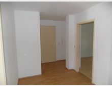 Weiß gestrichener Flur mit Laminatboden, einer beigen Eingangstür im Hintergrund und einem offenen Durchgang zu einem angrenzenden Raum auf der rechten Seite. An der Wand neben der Tür befindet sich eine Gegensprechanlage.