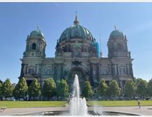 Historische Kathedrale mit grünen Kuppeln und reich verzierten Fassadenelementen, umgeben von Bäumen und Grünflächen; im Vordergrund ein Springbrunnen. Im Hintergrund ist der blaue Himmel sowie ein Teil eines Fernsehturms sichtbar.