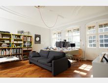 Helles Arbeitszimmer mit Holzfußboden, großen Sprossenfenstern und Stuckdecke. Im Raum befinden sich ein graues Sofa, ein Bücherregal, ein Schreibtisch mit mehreren Monitoren und ein kleiner Tisch.