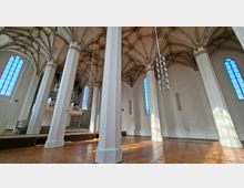 Innenraum einer hohen gotischen Hallenkirche mit weißen Pfeilern, Kreuzrippengewölbe und großen, schmalen Rundbogenfenstern. Im Hintergrund ist eine Orgel auf einer Empore zu sehen, von der Decke hängen mehrere moderne Leuchter über dem Holzfußboden.