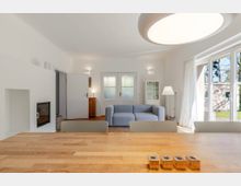 Helles, modern eingerichtetes Wohnzimmer mit Holztisch im Vordergrund, grauem Sofa, eingebautem Kamin und weißer Deckenleuchte. Große Fensterfront mit Vorhängen und Terrassentür lässt Tageslicht herein und bietet Blick ins Grüne.