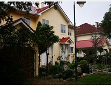 Zwei zweistöckige Einfamilienhäuser mit gelber Fassade und roten Satteldächern, umgeben von Gartenlandschaft mit Pflanzen, Gehwegen und Steinbeeten. Eine Laterne steht im Vordergrund, und ein Carport ist neben einem der Häuser sichtbar.