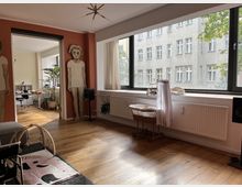 Wohnzimmer in einer modernen Wohnung mit Holzboden, großen Fenstern und Blick auf ein gegenüberliegendes Gebäude. Im Raum befinden sich ein Babyspielgerät, ein Kinderbett auf Rädern, Wandkunst in Form von großen Figuren sowie ein Durchgang zu einem Arbeitsbereich mit Schreibtisch und Pflanzen.