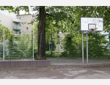 Ein kleiner Basketballplatz mit einem Metallkorb ohne Netz, umgeben von grünem Gitterzaun, Bäumen und dichtem Grün. Im Hintergrund sind Wohngebäude mit Balkonen zu sehen.