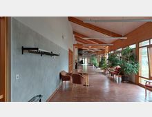 Ein heller Korridor mit großen Fensterfronten auf der rechten Seite, die viel Tageslicht hereinlassen; links eine graue Wand mit einer schwarzen Garderobe, daneben mehrere Türen aus Holz. Im Raum stehen Pflanzen in Töpfen sowie Sessel und Tische auf einem Boden aus Terrakottafliesen.