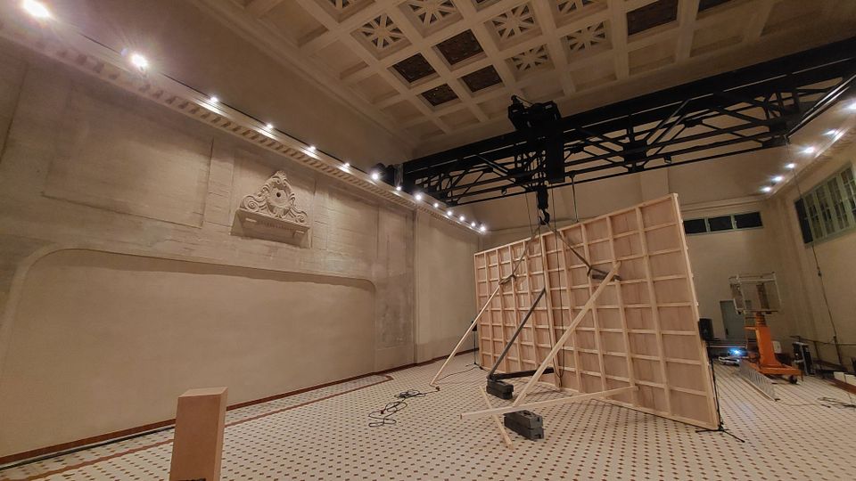 Großer Raum mit hoher Decke, verziert mit rechteckigen und durchbrochenen Mustern. Eine helle Wand mit einem kleinen Relief aus Stuck und eine Konstruktion aus Holzrahmen in der Raummitte; im Hintergrund sind moderne Beleuchtungselemente und technische Geräte zu sehen.