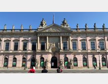 Barockgebäude mit rosafarbener Fassade und symmetrischem Design, gekrönt von zahlreichen Statuen. Der Mittelteil ist mit Säulen und einem reich verzierten Giebel gestaltet, darunter ein Wappenrelief; im Vordergrund Gehweg und Fußgänger.