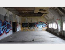Ein leerer, heruntergekommener Raum mit sichtbaren Schäden an Wänden und Decke, bedeckt mit Graffiti in unterschiedlichen Farben. Große, rechteckige Fenster auf der rechten Seite lassen Licht hinein, während der Boden aus ungleichmäßigen Platten besteht.
