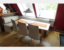 Essbereich in einem Dachgeschoss mit Holzboden, roter Wand und großem Tisch aus Holz, der von weißen Polsterstühlen und einer Eckbank umgeben ist. Drei große Dachfenster lassen natürliches Licht herein, und eine Hängelampe befindet sich über dem Tisch.