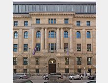 Historisches vierstöckiges Gebäude mit symmetrischer Sandsteinfassade, großen Fenstern und Reihen aus Säulen in der Mitte; im Erdgeschoss eine abgerundete Eingangstür. Autos und Fahrräder parken entlang der Straße davor.