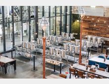 Innenansicht einer modernen Brauerei mit glänzenden Edelstahltanks auf erhöhten Plattformen, umgeben von Metallketten. Im Vordergrund befinden sich Sitzbereiche mit Holztischen und Stühlen sowie dekorative Lampen, die eine industrielle Atmosphäre schaffen.