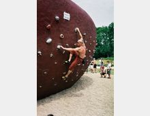 Ein muskulöser Mann klettert auf einer großen, roten Boulderwand mit künstlichen Griffen in einem Outdoor-Bereich. Im Hintergrund befinden sich andere Personen auf einem Kiesplatz sowie grüne Bäume.