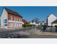 Straßenszene mit einem zweigeschossigen Gebäude aus rotem Backstein und weißen Fensterrahmen auf der linken Seite, beschriftet mit „HO Obst und Gemüse“. Ein Straßenschild zeigt „Hermann-Löns-Straße“, daneben Geh- und Radwegschilder. Im Hintergrund sind weitere Wohnhäuser und ein kleiner Platz mit minimaler Begrünung.