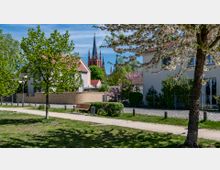 Ein gepflegter Weg in einer Wohngegend mit grünen Rasenflächen und blühenden Bäumen im Vordergrund, flankiert von modernen Gebäuden mit roten Dächern; im Hintergrund ist eine Kirche mit hohen, spitzen Türmen sichtbar.