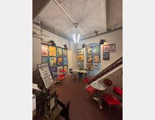 Kleiner Innenraum mit weiß gestrichenen Ziegelwänden, dekoriert mit bunten Plakaten, die Tiere und Zoothemen zeigen. Der Raum verfügt über Metalltische mit roten und blauen Stühlen, eine rötlich-braune Bodenfläche und eine auffällige Hängelampe unter der hohen Decke.