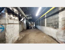 Ein langer, schmaler Industriegang mit abgenutzten weißen Wänden, sichtbaren Rohren und Kabeln an der Decke. Auf der rechten Seite befinden sich Fenster mit geteilter Glasfläche, die Licht einlassen; der Boden ist uneben und weist Wasserpfützen auf.