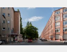 Eine städtische Straßenecke mit einem kleinen Imbiss (&quot;Mini Pizza&quot;) im Erdgeschoss eines schlichten Wohnhauses links und einem langen, rot-weißen Backsteingebäude rechts. In der Mitte verläuft eine von Bäumen gesäumte gepflasterte Straße, an der mehrere parkende Autos stehen.