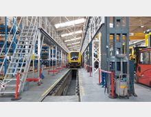 Innenansicht einer Werkstatthalle für Züge mit hoher Decke und Oberlichtern. Im Zentrum steht ein gelber Zug über einem Wartungsgraben, umgeben von Arbeitsplattformen, Werkzeugen und Regalen voller Ersatzteile.