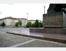 Offener Platz mit gepflastertem Boden, flankiert von einem großen Denkmal mit der Inschrift "ROT FRONT" auf einem bronzefarbenen Sockel. Im Hintergrund befinden sich mehrstöckige Wohngebäude mit hellen Fassaden und roten Dächern sowie vereinzelte Bäume und Straßenbeleuchtung.