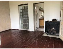 Ein einfacher Raum mit dunkelbraunem Holzboden, einer Glasbausteinwand und einem offenen Durchgang zu einer kleinen Küche im Hintergrund. Rechts steht ein schwarzer Kaminofen mit Zubehör, die Wände sind schlicht in Weiß und Beige gehalten.