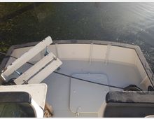 Bug eines kleinen Bootes mit weißer Innenverkleidung und abblätternder Farbe, ausgestattet mit einer schlichten Sitzbank. Im Hintergrund ist ruhiges Wasser mit schwimmender grüner Vegetation zu sehen.