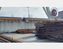 Industriegelände mit gestapelten Metallträgern im Vordergrund, einer Betonschutzwand in der Mitte und Güterwaggons im Hintergrund. Ein großes rot-weißes Seilrad steht rechts neben einer Halle im industriellen Baustil.