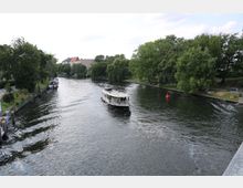 Ein Fluss mit einem weißen Ausflugsboot in der Mitte, umgeben von grünen Bäumen und Parkanlagen auf beiden Uferseiten. Im Hintergrund sind einige Gebäude sichtbar, während sich auf der rechten Flussseite Personen im Park entspannen.