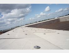 Flaches Dach mit einer Oberfläche aus hellen Platten, die mit Schrauben befestigt sind, daneben eine Holzplattform. Im Hintergrund sind eine städtische Umgebung mit Gebäuden, Bäumen und ein bewölkter Himmel zu sehen.