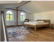 Schlafzimmer in einem Dachgeschoss mit schräger Decke, Holzbalken und zwei halbrunden Fenstern. Der Raum ist mit einem Holzbett, einem bunten Teppich, einem kleinen Beistelltisch und einem Sessel eingerichtet.