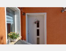 Eingangsbereich eines Wohnhauses mit orangefarbener Putzfassade, weißer Haustür mit drei schmalen Glasfeldern und kleinem Türkranz. Links daneben ein großes Fenster mit Blumenkasten auf der Fensterbank sowie eine Außenwandleuchte.