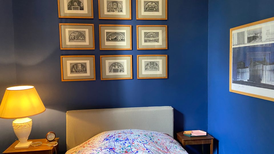Schlafzimmer mit dunkelblauen Wänden und einem Bett mit hellgrauem Kopfteil sowie einer bunt gesprenkelten Bettdecke. Über dem Bett hängen neun gerahmte Schwarzweiß-Drucke in einem Raster; daneben stehen zwei Nachttische, einer mit Tischlampe und kleinem Wecker.