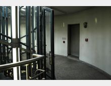Treppenhaus mit einer Wendeltreppe aus Metall und Glaswänden auf der linken Seite, sowie einem glatten grauen Boden. An der rechten Wand befinden sich eine Tür, ein Rauchabzugsschalter und eine Feuerlöschvorrichtung.