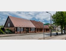 Straßenszene mit einem einstöckigen Wohnhaus im Fachwerkstil mit Backsteinmauern und roten Dachziegeln, an einem mit Kopfsteinpflaster belegten Weg. Rechts neben dem Haus stehen Bäume und eine Straßenlaterne, im Hintergrund weitere Wohnhäuser.