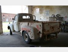 Ein alter, rostiger Pickup-Truck steht in einer offenen Garage oder Werkstatt mit Betonboden. An den Wänden hängen Reparaturwerkzeuge, Schilder und Autoteile, während draußen fragmentarisch alte Backsteingebäude und ein grünes Feld sichtbar sind.