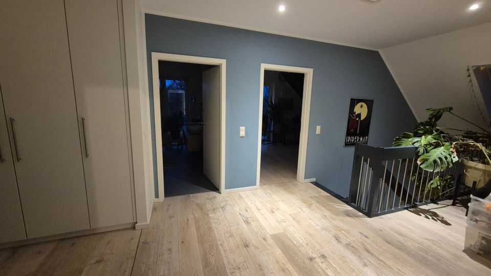 Flur in einem modernen Wohnhaus mit Holzfußboden, blauer Wandfarbe und drei Türen, die zu angrenzenden Räumen führen. Links befindet sich ein großer, weißer Einbauschrank, und rechts ist eine Treppe mit schwarzem Geländer sowie eine Zimmerpflanze sichtbar.