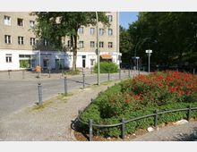 Ein kleiner Platz mit gepflasterten Wegen, umgeben von niedrigen Metallpollern. Im Vordergrund ein mit roten Blumen bepflanztes Beet, im Hintergrund ein mehrstöckiges Wohngebäude mit Geschäften im Erdgeschoss.