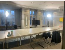 Tagungsraum mit rechteckigem Konferenztisch und modernen Stühlen. Große Fenster lassen Tageslicht herein, während Studiolichter und eine Kamera auf Stativen aufgestellt sind; zusätzlich sind ein Fernseher und Desinfektionsmittel auf dem Tisch zu sehen.