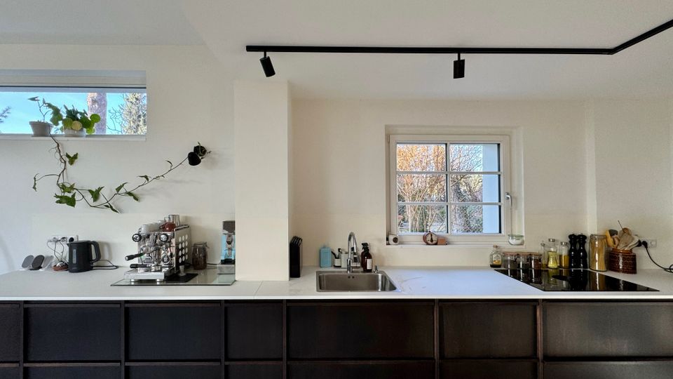 Moderne Küche mit langen dunklen Unterschränken und weißer Arbeitsplatte, mittigem Spülbecken und zwei Fenstern mit Blick auf Bäume. Auf der Arbeitsfläche stehen Kaffeemaschine, Wasserkocher, Gewürzgläser und Öle; an der Decke verläuft eine schwarze Schienenbeleuchtung.